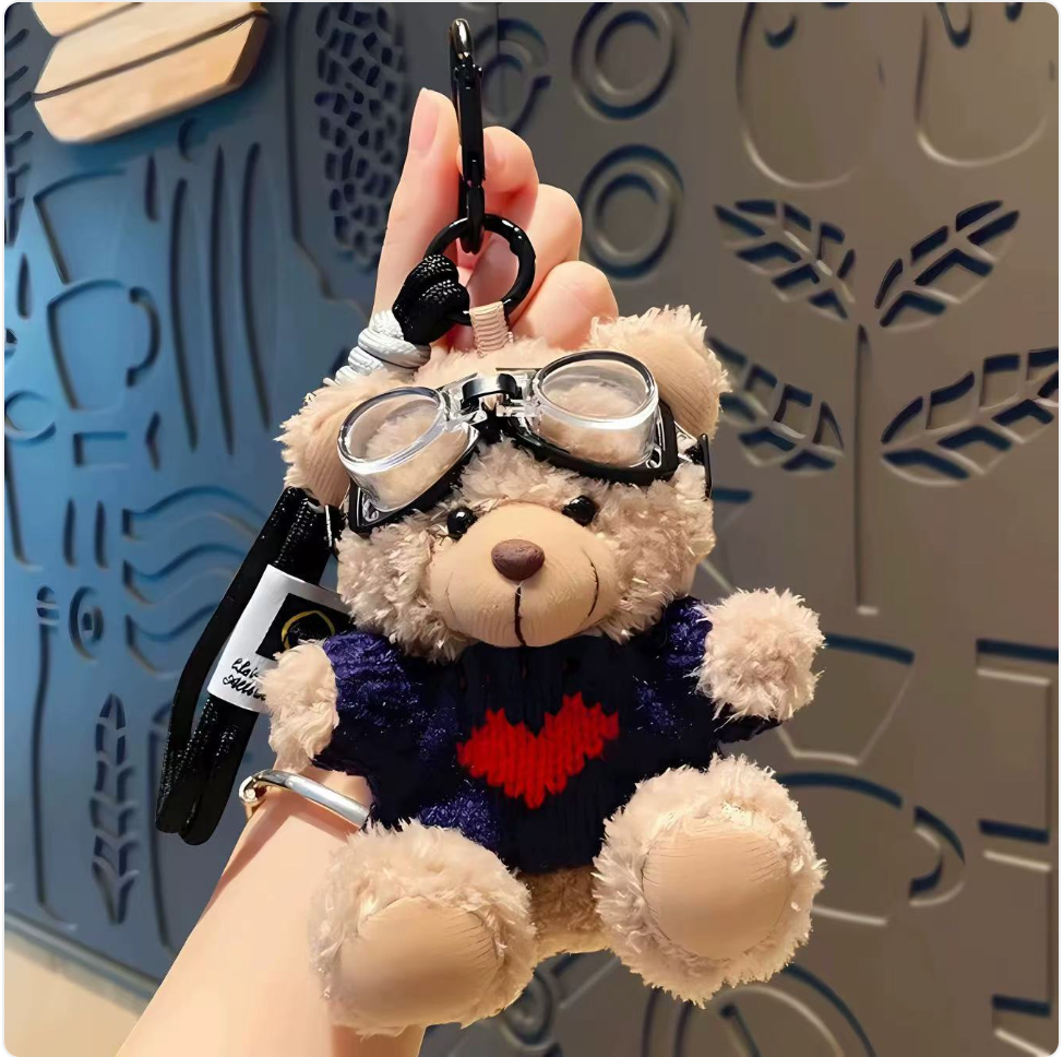 Teddy bear keychain with goggles held by a hand against a decorative wall.
